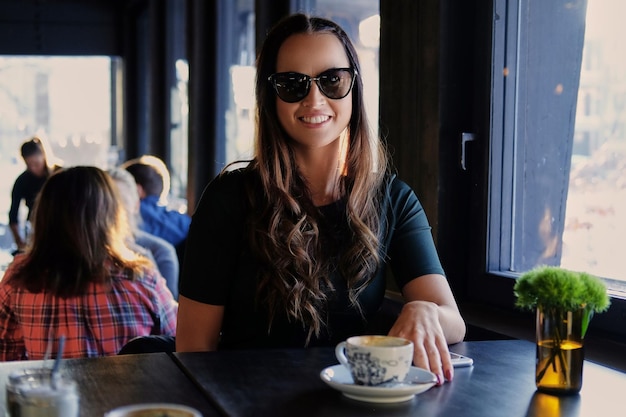 Ritratto di donna bruna sorridente in occhiali da sole, beve il caffè del mattino in un bar.