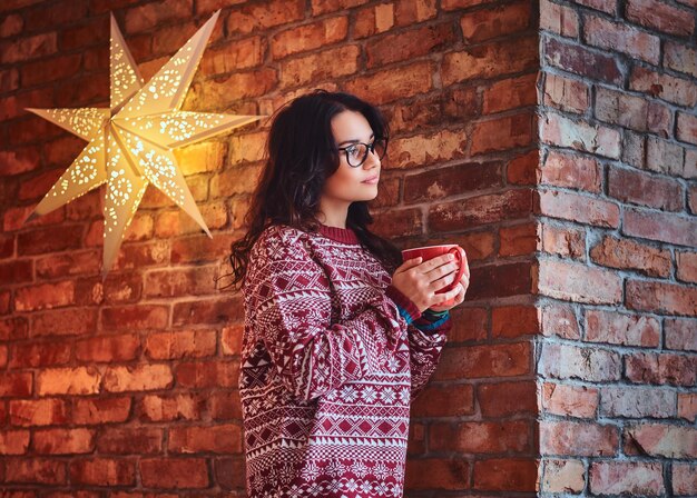 Ritratto di donna bruna vestita con un maglione rosso beve caffè sopra il muro di un mattone.