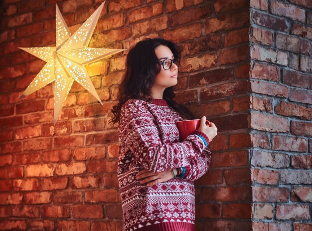 Ritratto di donna bruna vestita con un maglione rosso beve caffè sopra il muro di un mattone.