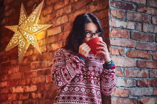 Ritratto di donna bruna vestita con un maglione rosso beve caffè sopra il muro di un mattone.