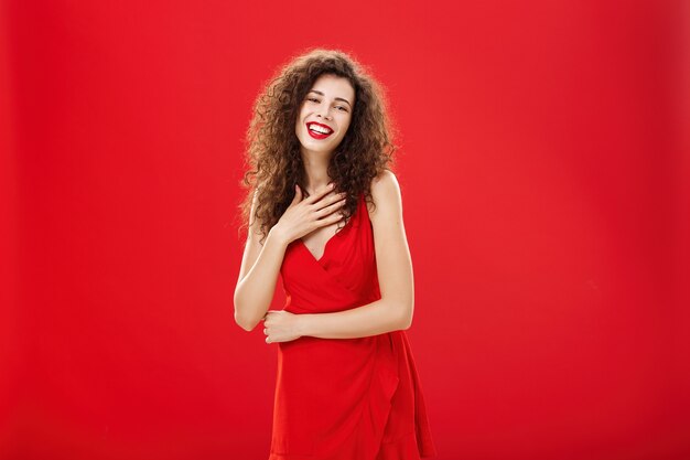 Ritratto di donna caucasica affascinante felice e grata elegante con l'acconciatura riccia che tiene la palma sul petto che sorride e che ride divertito posa in abito da sera elegante su sfondo rosso.