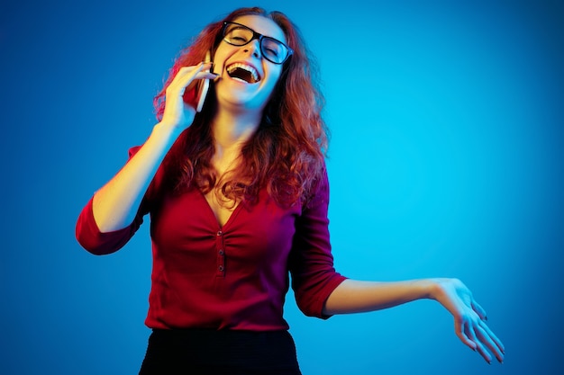 Ritratto di donna caucasica isolato su sfondo blu studio in luce al neon. Bellissimo modello femminile con i capelli rossi in casual. Concetto di emozioni umane, espressione facciale, vendite, annuncio. Parlando al telefono.