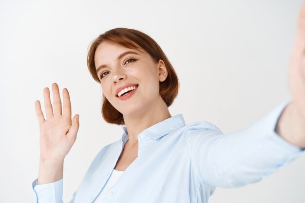 Ritratto di donna che agita la mano per salutare in video chat, tenendo lo smartphone in mano tesa, salutando un amico, in piedi contro il muro bianco