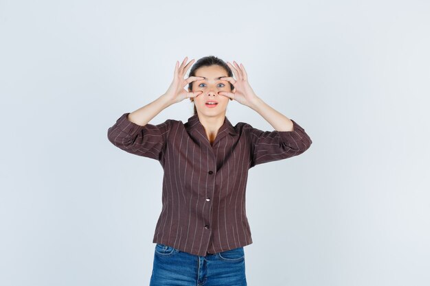 Ritratto di donna che apre gli occhi con le dita in camicia, jeans e guarda perplesso vista frontale