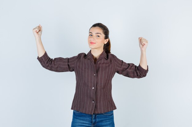 Ritratto di donna che mostra il gesto del vincitore in camicia, jeans e guarda con orgoglio la vista frontale