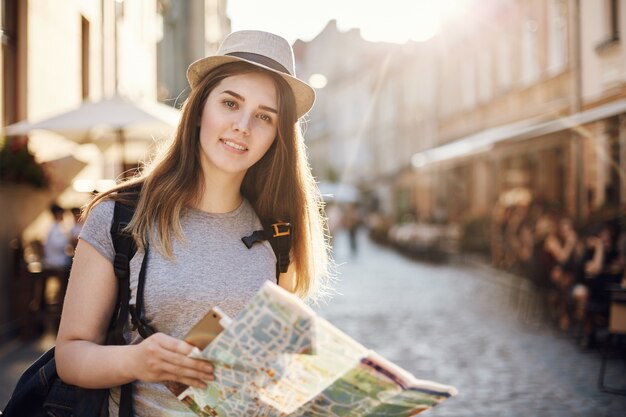 Ritratto di donna che viaggia per il mondo utilizzando una mappa e tablet, in piedi in una piccola città europea che guarda l'obbiettivo.