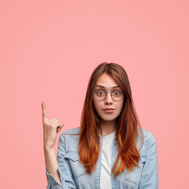 Ritratto di donna con la pelle lentigginosa e lunghi capelli lisci, solleva l'indice verso l'alto, vestito con una giacca di jeans, isolato su sfondo rosa