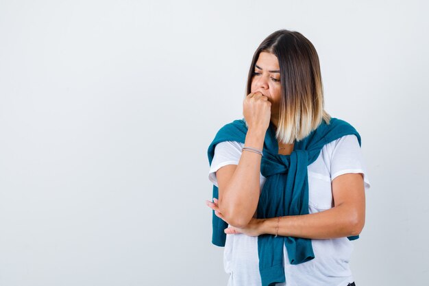 Ritratto di donna con maglione legato che morde il pugno emotivamente con una maglietta bianca e guarda una vista frontale pensierosa