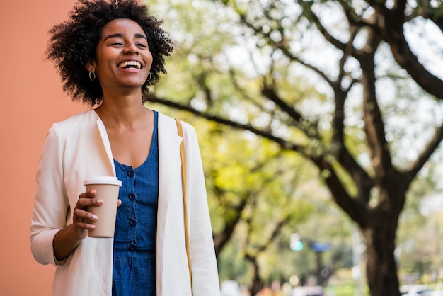 Ritratto di donna d'affari afro sorridente e tenendo una tazza di caffè mentre si trovava all'aperto sulla strada. Business e concetto urbano.