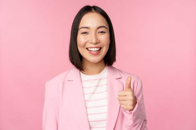Ritratto di donna d'affari asiatica che sorride soddisfatta mostrando i pollici in su elogia e approva in piedi in tuta su sfondo rosa