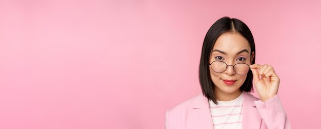 Ritratto di donna d'affari asiatica con gli occhiali che guarda incuriosito la fotocamera e sorridente commessa professionista che fissa con interesse sfondo rosa