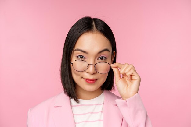Ritratto di donna d'affari asiatica con gli occhiali che guarda incuriosito la fotocamera e sorridente commessa professionista che fissa con interesse sfondo rosa