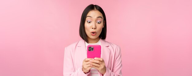 Ritratto di donna d'affari asiatica con la faccia sorpresa che utilizza l'app per smartphone che indossa un tailleur Ragazza coreana con il telefono cellulare e l'espressione del viso eccitata sfondo rosa