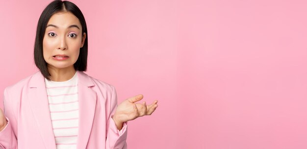 Ritratto di donna d'affari asiatica confusa che scrolla le spalle le spalle guardando all'oscuro e perplessa non so non posso dire in piedi su sfondo rosa in tuta da ufficio