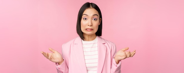Ritratto di donna d'affari asiatica confusa che scrolla le spalle le spalle guardando all'oscuro e perplessa non so non posso dire in piedi su sfondo rosa in tuta da ufficio