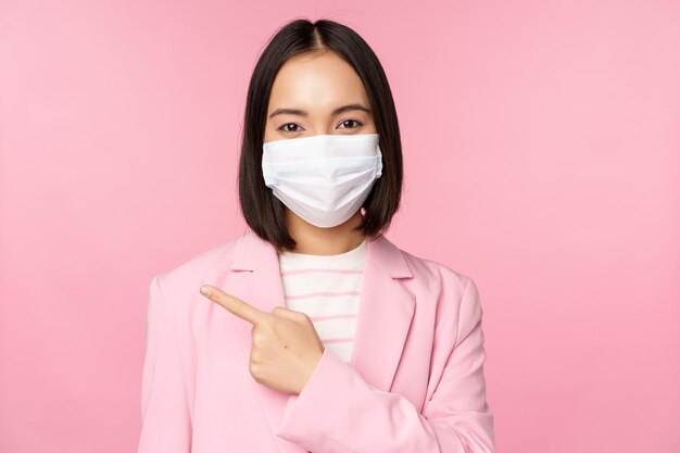 Ritratto di donna d'affari asiatica in maschera medica e tuta che punta il dito a sinistra che mostra lo sfondo rosa dello studio dell'insegna della società pubblicitaria