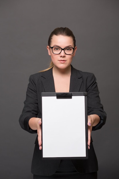 Ritratto di donna d'affari seria in possesso di una clip board davanti a lei con una pagina vuota. Bella signora con gli occhiali in posa in tailleur nero.