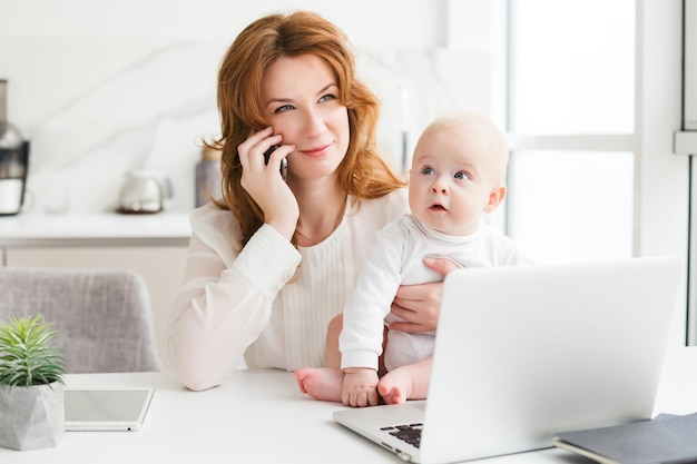 Ritratto di donna d'affari sorridente seduta al tavolo con il laptop e parlando sul suo cellulare mentre tiene in mano il suo piccolo bambino carino