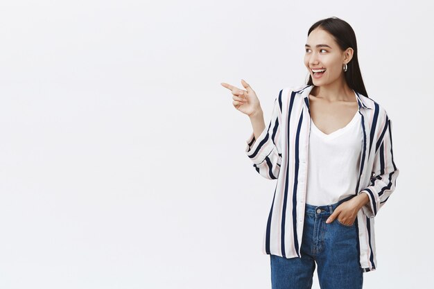 Ritratto di donna di bell'aspetto amichevole in jeans e camicetta a righe alla moda, tenendo la mano in tasca, indicando e guardando a sinistra con un ampio sorriso