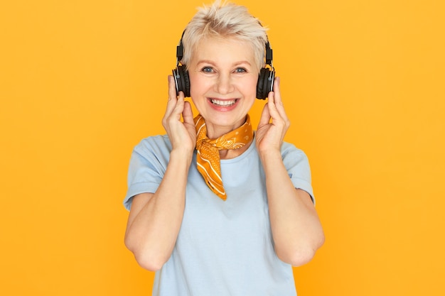 Ritratto di donna di mezza età attraente allegra con i capelli corti tinti che ascolta le canzoni usando le cuffie senza fili, avendo un'espressione facciale gioiosa.