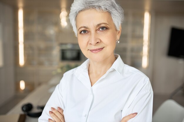 Ritratto di donna di mezza età seria fiduciosa con i capelli corti grigi, occhi verdi, rughe e sorriso affascinante in posa all'interno con le braccia conserte