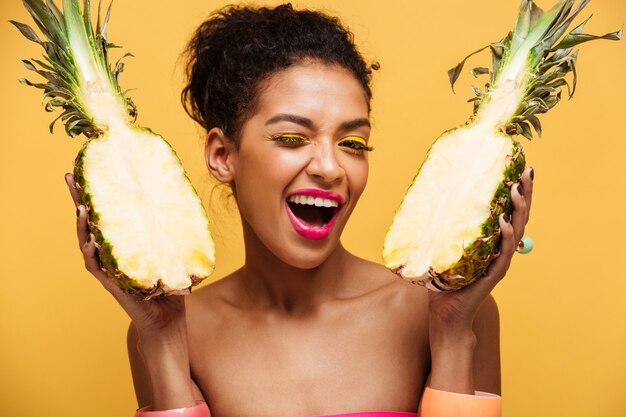 Ritratto di donna di razza mista felice con aspetto moda tenendo spaccata di ananas fresco a metà isolato, sopra la parete gialla