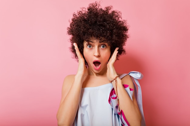 Ritratto di donna divertente con capelli ricci corti pone sul rosa con la bocca aperta e tiene le mani vicino al viso con brillanti emozioni sorprese