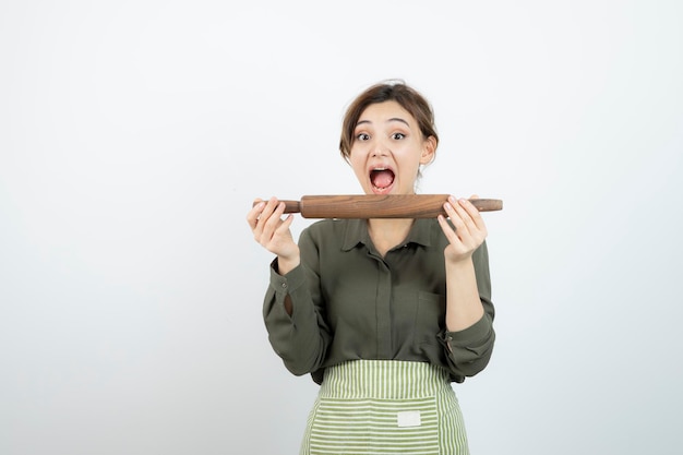 Ritratto di donna divertente in grembiule che cerca di mordere un mattarello di legno. Foto di alta qualità