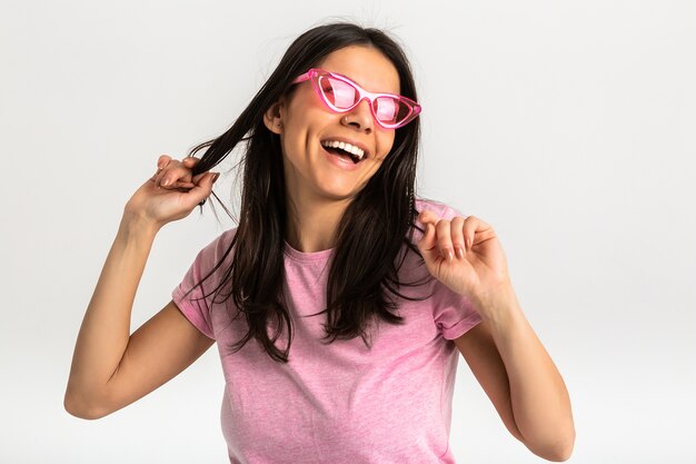 Ritratto di donna emotiva abbastanza sorridente in camicia rosa e occhiali da sole alla moda, denti bianchi, posa positiva isolata