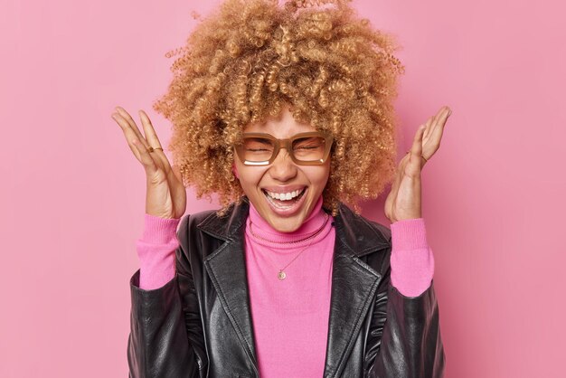 Ritratto di donna emozionale dai capelli ricci esclama di gioia tiene i palmi alzati si sente pazzo indossa una giacca di pelle nera alla moda isolata su sfondo rosa felice di ricevere il regalo di compleanno