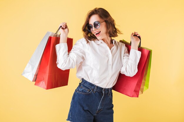 Ritratto di donna felice in occhiali da sole in piedi con borse della spesa colorate in mano su sfondo rosa. Giovane donna in piedi in camicia bianca e pantaloncini di jeans