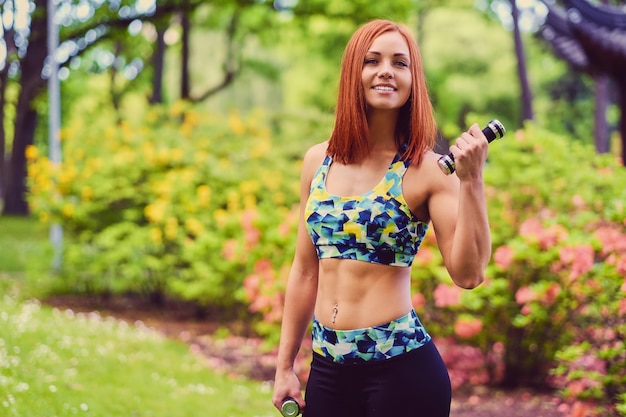 Ritratto di donna fitness rossa tiene manubri in un parco all'aperto.