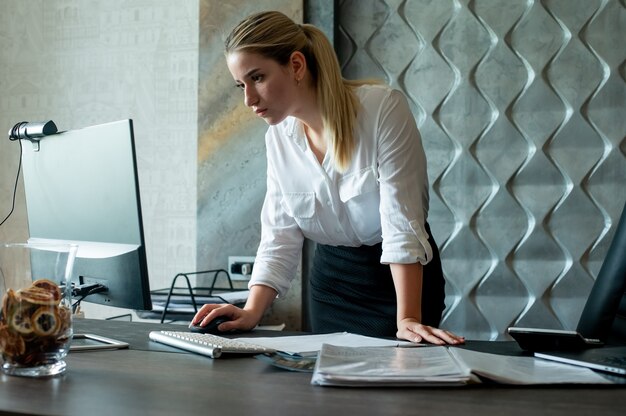 Ritratto di donna giovane lavoratore di ufficio in piedi alla scrivania in ufficio con documenti utilizzando il computer con espressione seria e fiduciosa sul viso lavorando in ufficio