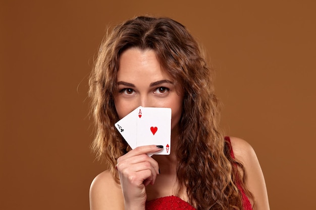Ritratto di donna giovane o dai capelli castani sorridente, con in mano un paio di assi che indossano un abito da cocktail rosso su sfondo marrone. Concetto di casinò, industria del gioco d'azzardo