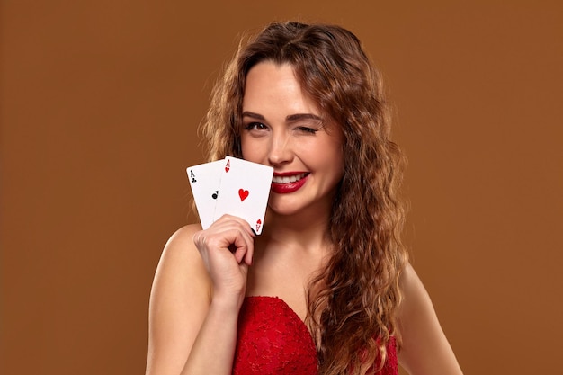 Ritratto di donna giovane o dai capelli castani sorridente, con in mano un paio di assi che indossano un abito da cocktail rosso su sfondo marrone. Concetto di casinò, industria del gioco d'azzardo