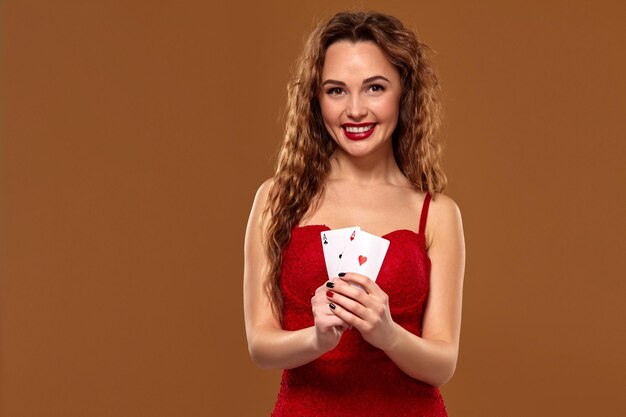 Ritratto di donna giovane o dai capelli castani sorridente, con in mano un paio di assi che indossano un abito da cocktail rosso su sfondo marrone. Concetto di casinò, industria del gioco d'azzardo