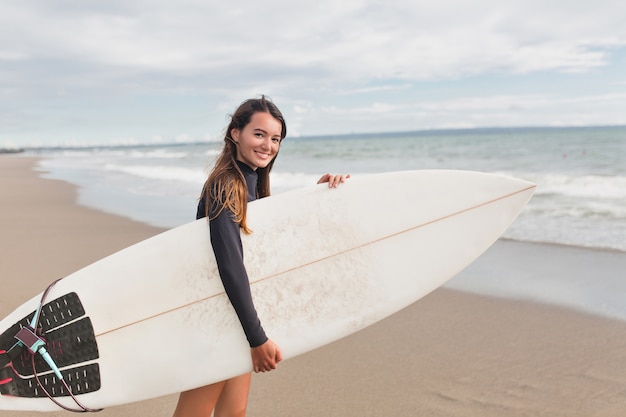 Ritratto di donna graziosa affascinante con i capelli lunghi che si prepara per una lezione di surf, sorride alla telecamera, tenendo la tavola da surf e si leva in piedi sulla riva dell'oceano. Buona giornata di sole, stile di vita attivo