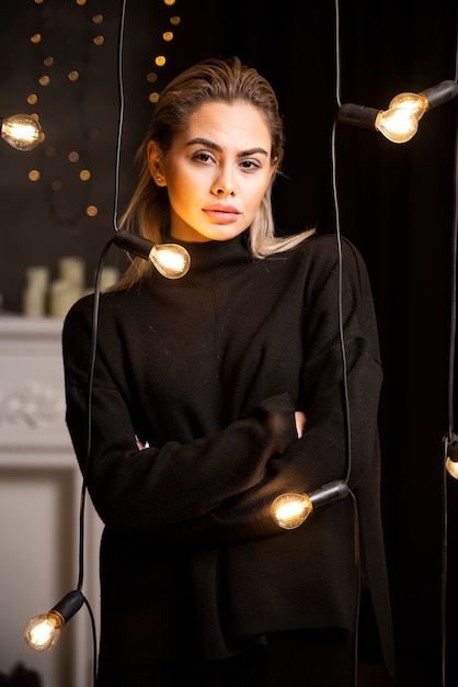 Ritratto di donna graziosa che posa in maglione scuro in piedi vicino alle lampade.