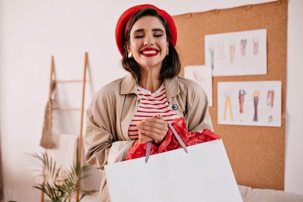 Ritratto di donna in cappello rosso e camicia a righe posa felicemente con la borsa della spesa. Ragazza carina con rossetto luminoso in berretto e cappotto beige sorride.