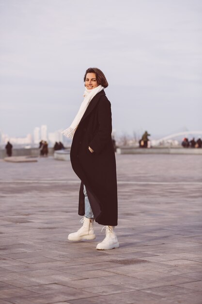 Ritratto di donna in cappotto invernale nero e sciarpa bianca sulla piazza della città.