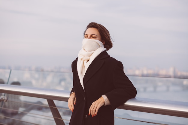 Ritratto di donna in cappotto invernale nero e sciarpa bianca sulla piazza della città.