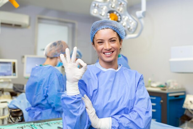 Ritratto di donna infermiere chirurgo O membro del personale vestito con camice chirurgico scrub maschera e retina per capelli nel teatro della sala operatoria dell'ospedale facendo contatto visivo sorridente mostrando segno ok