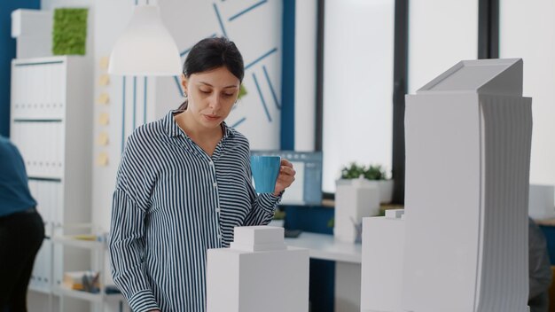 Ritratto di donna ingegnere guardando il modello di costruzione e bere una tazza di caffè al lavoro. Architetto che analizza la maquette per progettare la struttura e il layout della costruzione in uno studio di architettura.