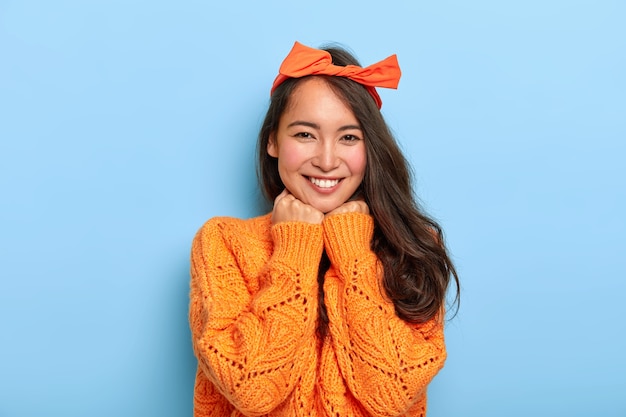 Ritratto di donna millenaria bruna felice tiene entrambe le mani sotto il mento, sorride piacevolmente, indossa la fascia arancione dell'arco, maglione lavorato a maglia arancione caldo