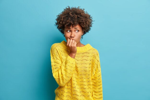 Ritratto di donna nervosa tiene le mani vicino alla bocca si sente preoccupato prima che un importante colloquio esiti su qualcosa vestito con un maglione giallo lavorato a maglia pone contro il muro blu dello studio