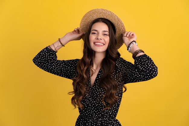 Ritratto di donna piuttosto attraente in posa isolato su sfondo giallo che indossa abito nero punteggiato e cappello di paglia alla moda boho tendenza moda primavera estate accessori stile sorridente umore felice