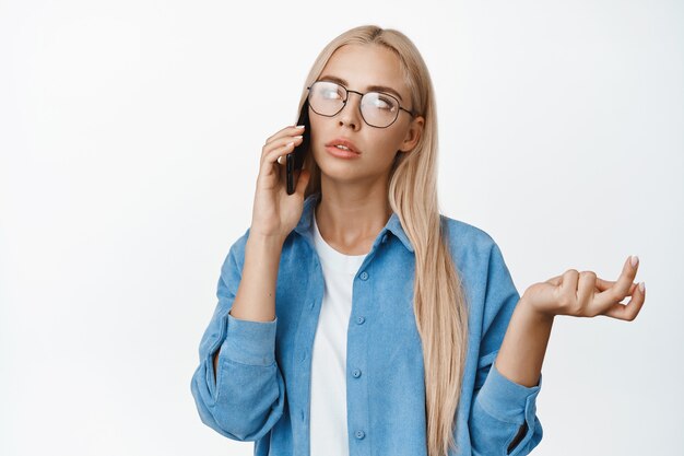 Ritratto di donna premurosa con gli occhiali che parla al telefono cellulare e scrolla le spalle, sembra perplessa durante una chiamata, in piedi su bianco