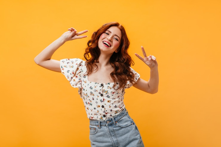 Foto gratuito ritratto di donna riccia in camicetta e gonna in denim che mostra il segno di pace su sfondo arancione.