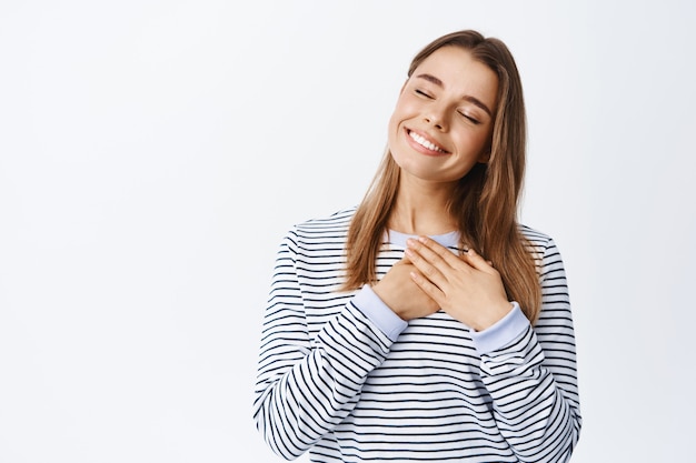 Ritratto di donna romantica felice chiude gli occhi, tenendosi per mano sul cuore e sognando ad occhi aperti, ricorda un bel ricordo, avendo sentimenti puri nel cuore, muro bianco