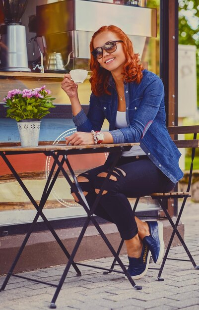 Ritratto di donna rossa in occhiali da sole, beve caffè in un bar in una strada.